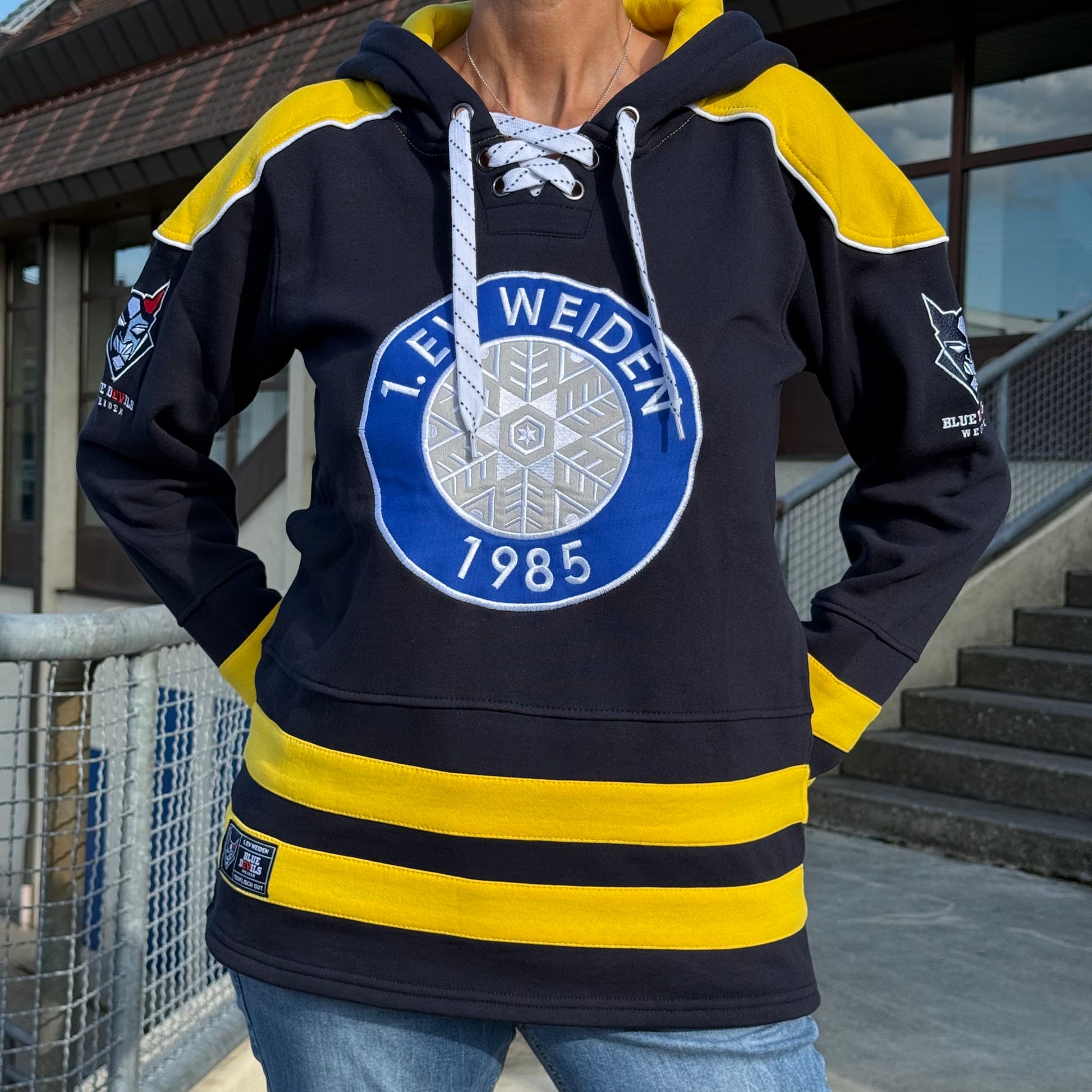 Person trägt einen dunkelblauen Kapuzenpullover mit gelben Akzenten und dem Logo des Eishockeyvereins 1. EV Weiden (gegründet 1985) auf der Brust.