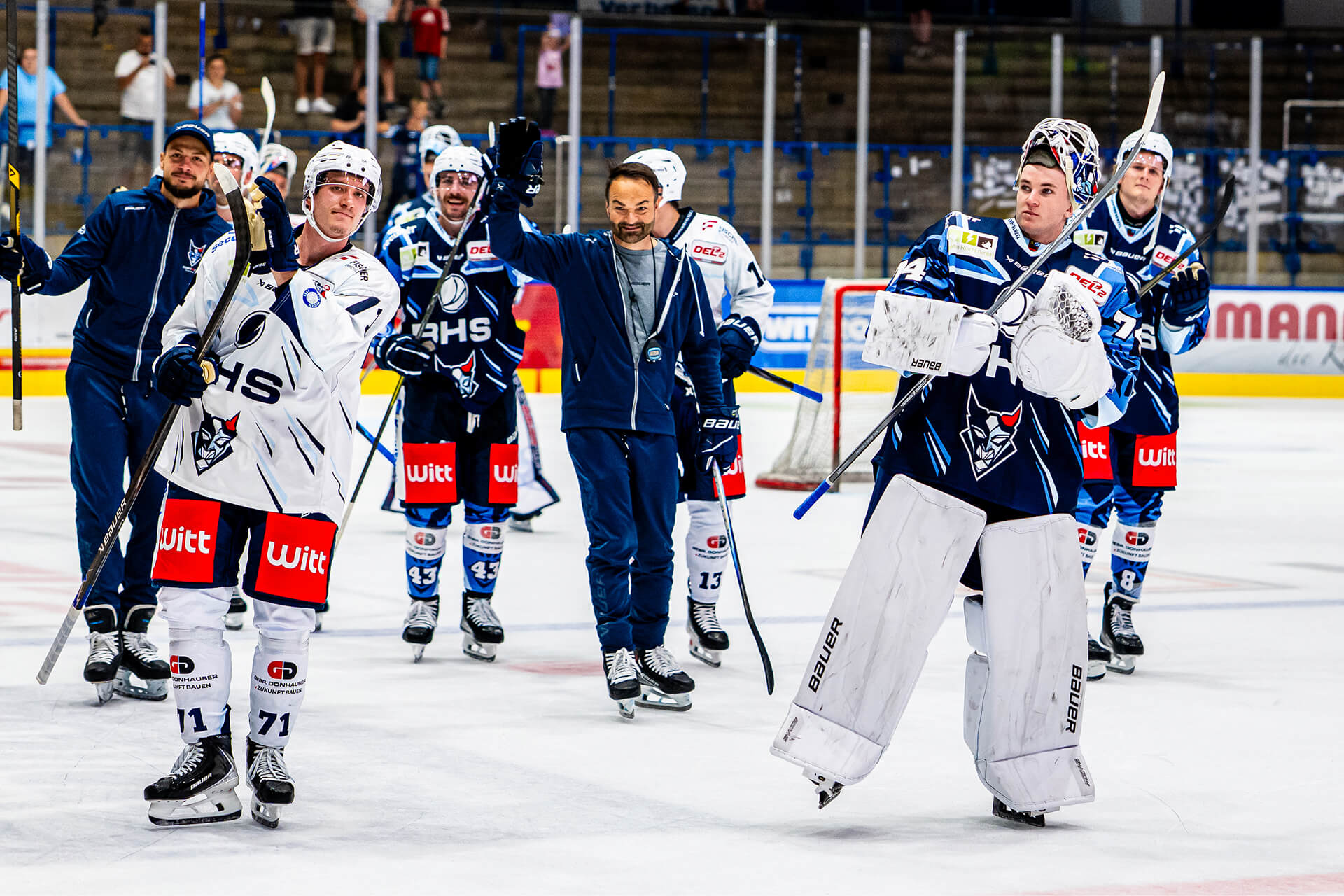 Eishockeyspieler und Trainer eines Teams winken und jubeln gemeinsam auf dem Eis nach einem Spiel. Einige tragen blaue, andere weiße Trikots, ein Torhüter in voller Ausrüstung steht vorne.